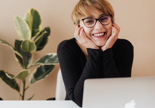 Empowered business woman smiling with hands around face sitting at a desk with an Apple computer and a leafy plant in the background.